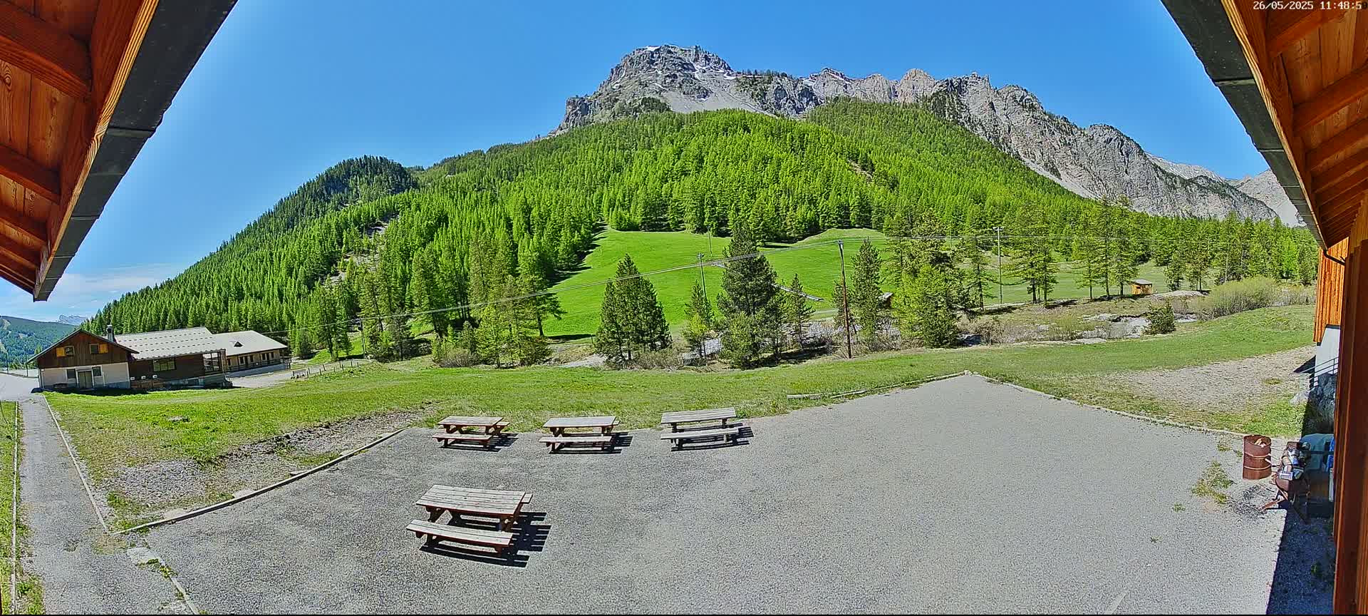 A gravel area with several picnic tables sits in front of a building, overlooking a lush green hillside dotted with trees and a mountainous backdrop under a clear blue sky.