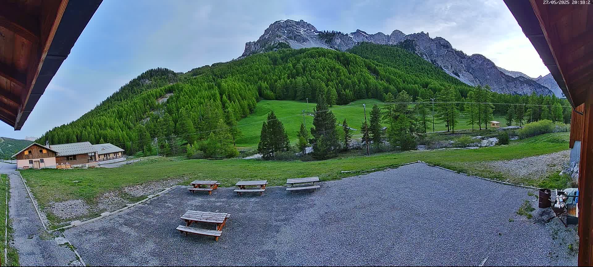 A gravel area with picnic tables sits in front of a wooden building, overlooking a verdant mountain valley under a partly cloudy sky.