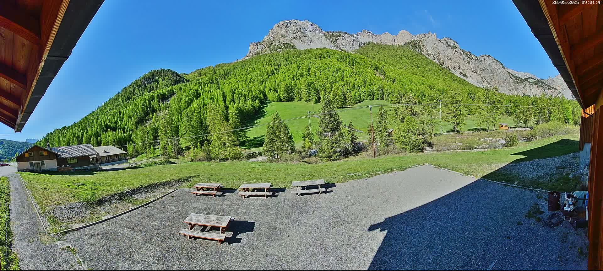 A gravel area with several picnic tables sits in front of a building, overlooking a lush green hillside dotted with trees and a rocky mountain under a clear blue sky.