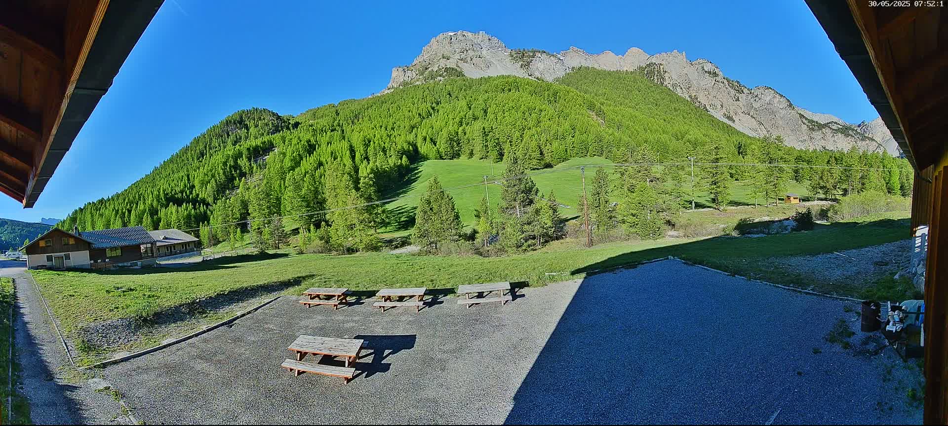 A gravel patio with several picnic tables sits before a building and a lush green mountainside under a clear blue sky.