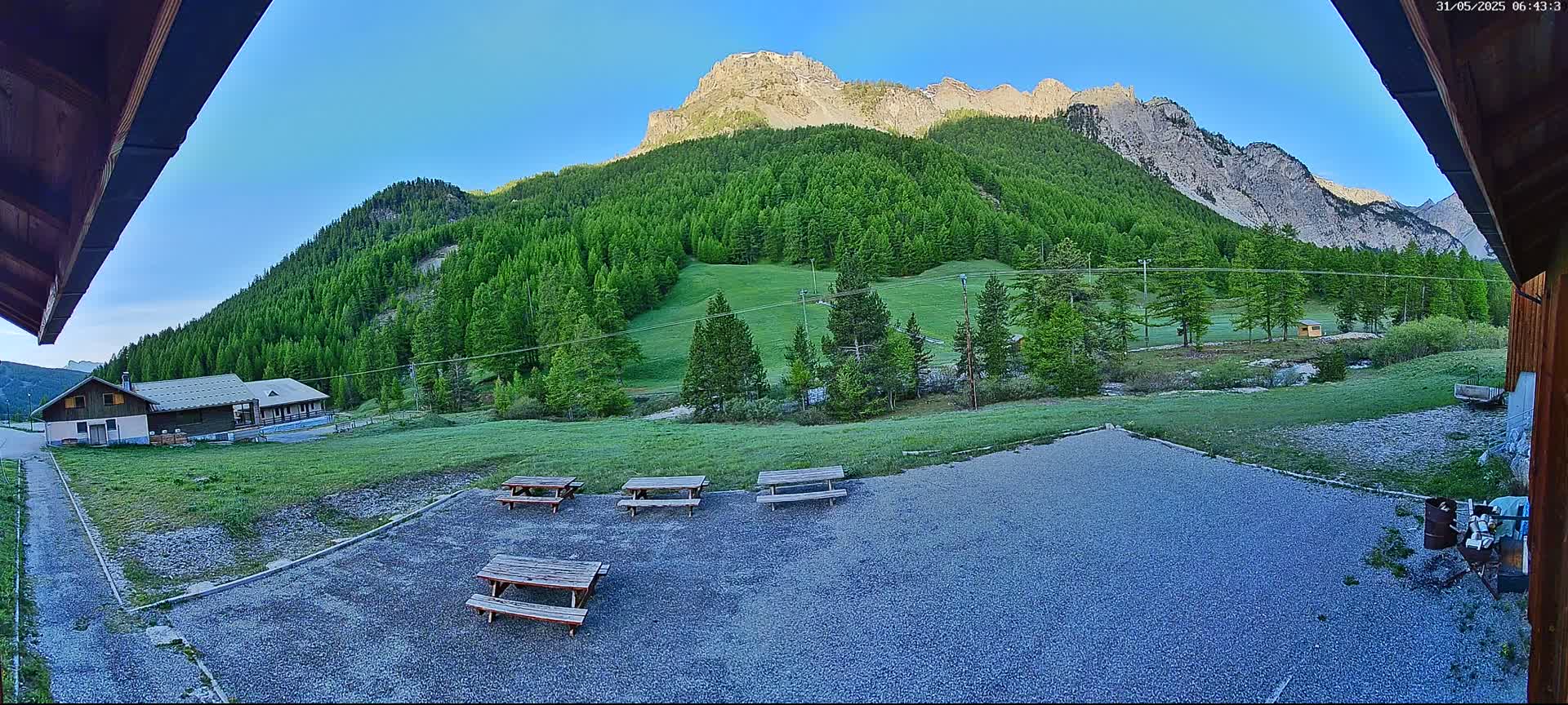 A gravel area with several picnic tables sits before a grassy field, a building, and a large, verdant mountain under a clear blue sky.