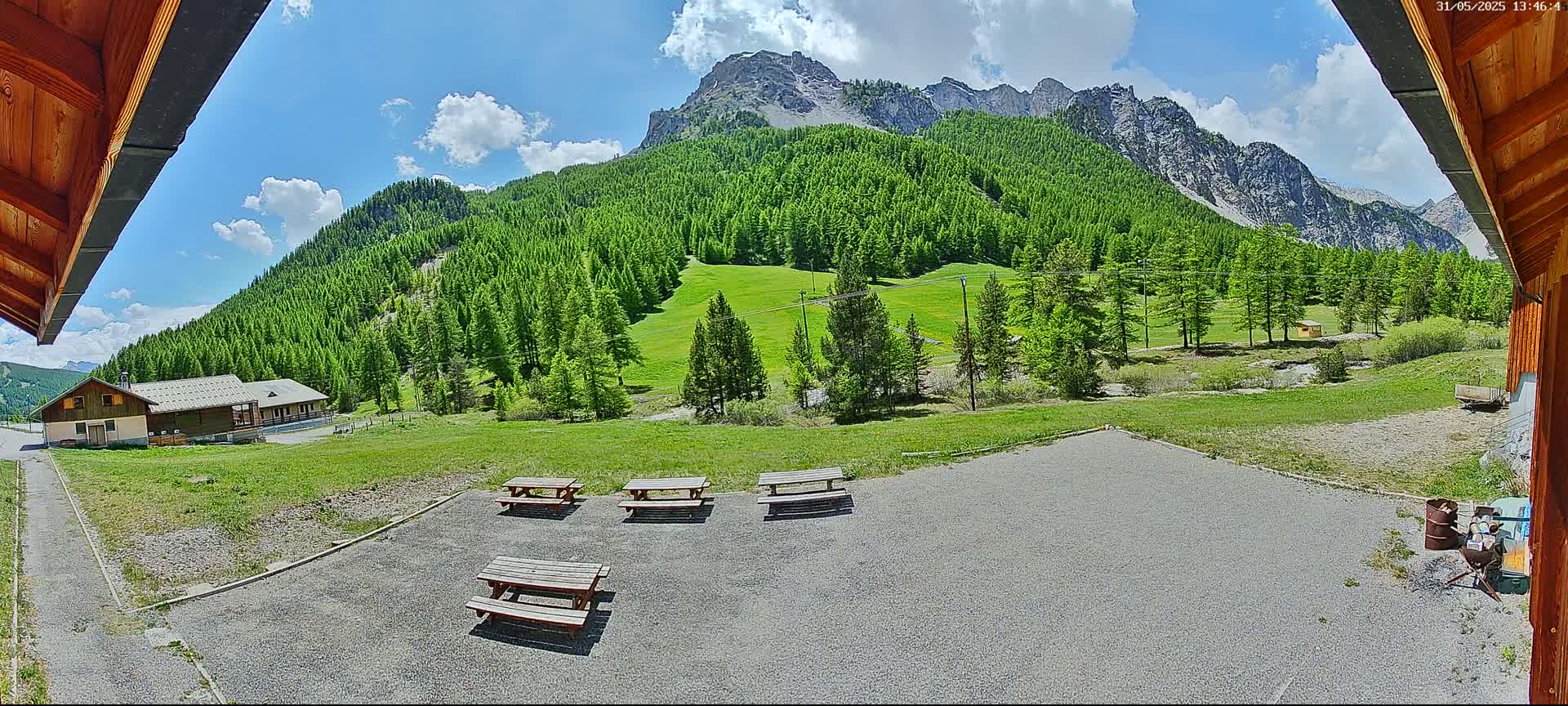 A sunny day reveals a view of a grassy area with several picnic tables, a building, and a mountain range covered in lush green trees.