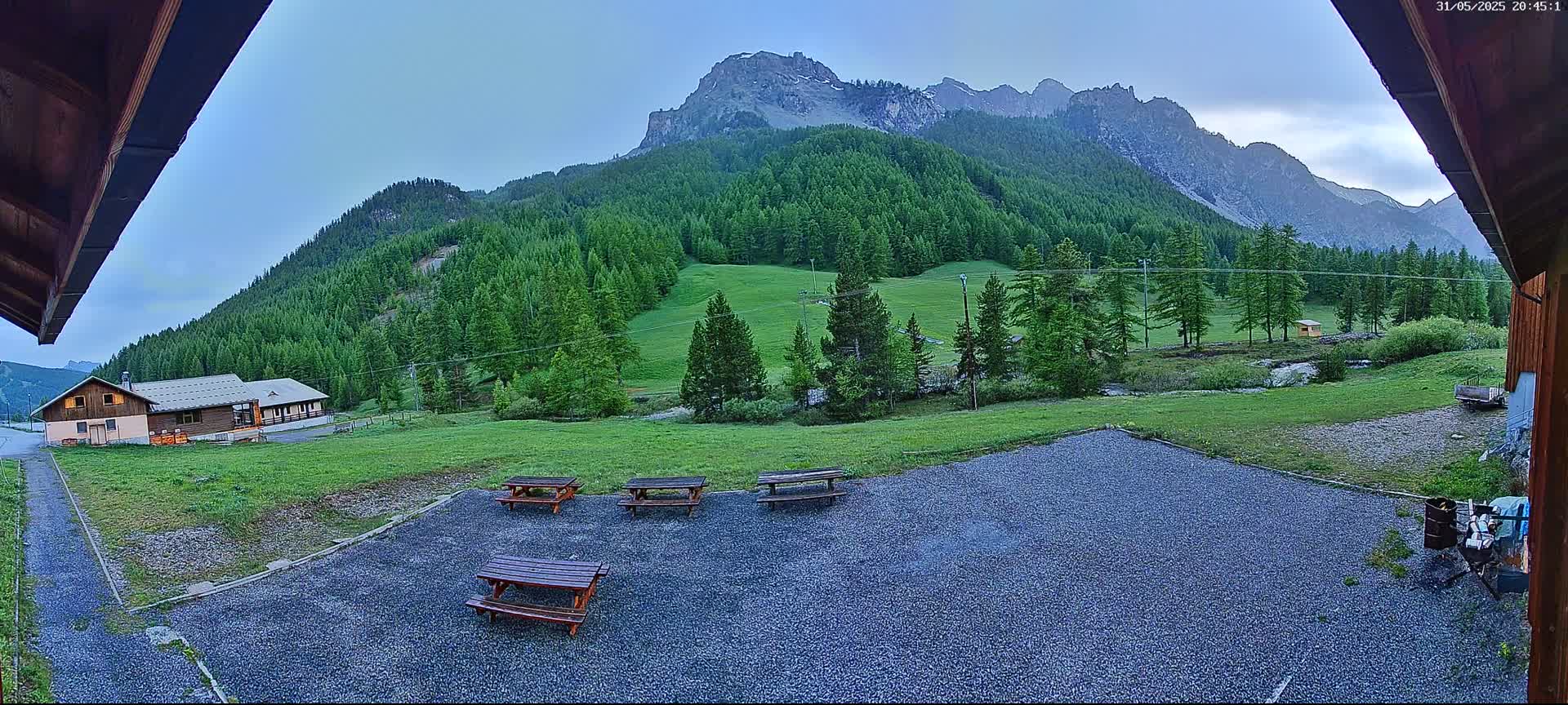 A gravel area with picnic tables sits in front of a building, overlooking a grassy field, a forest of evergreen trees, and a mountain range under a clear, twilight sky.