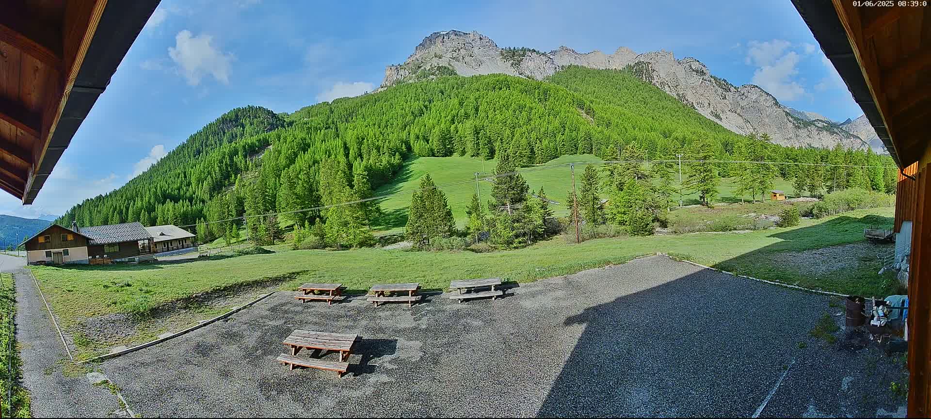 A sunny day reveals a gravel area with picnic tables overlooking a lush green valley and mountains in the background.