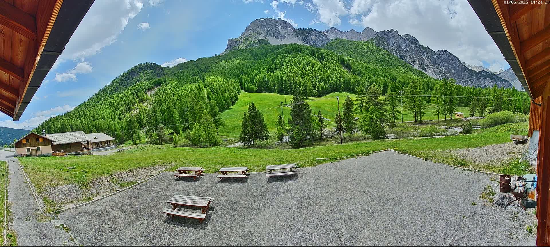 A gravel area with several picnic tables sits in front of a building, overlooking a grassy valley and a mountain range under a partly cloudy sky.