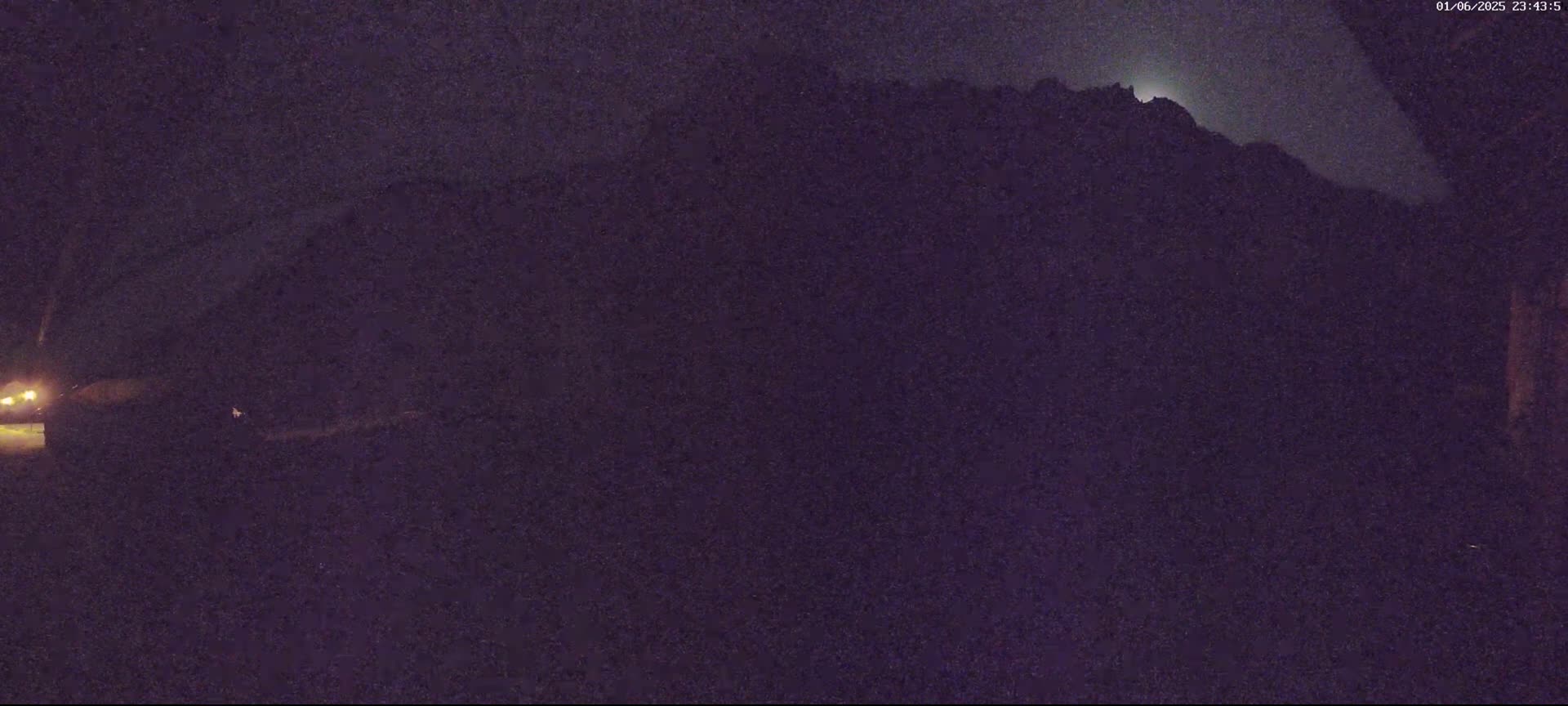 A nighttime, low-light image shows a dark, silhouetted mountain range under a mostly clear sky with a bright moon or planet visible.