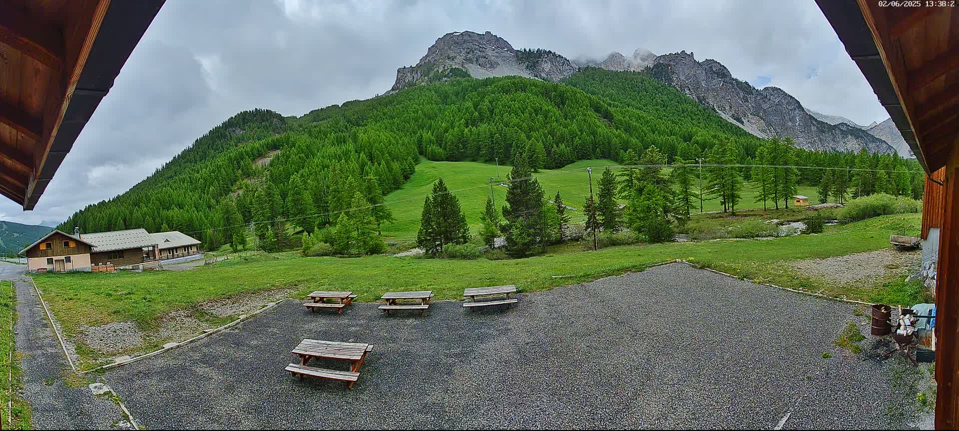 A gravel area with picnic tables sits before a grassy field and a mountain range covered in lush green trees under a cloudy sky.