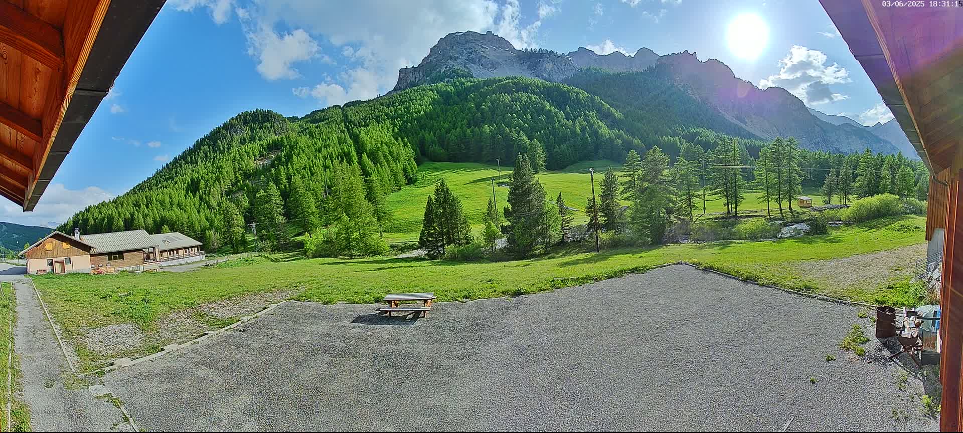 A sunny day in the mountains shows a gravel area with a picnic table, a grassy field, a building, and a mountain range covered in lush green trees.