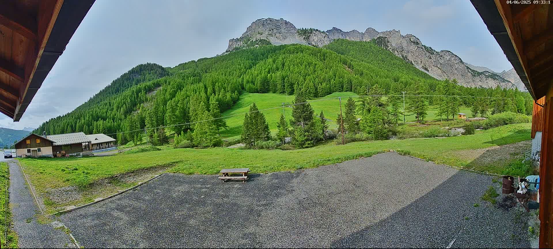 A gravel area with a picnic table sits in front of a building, overlooking a lush green valley and a mountainous backdrop under a partly cloudy sky.