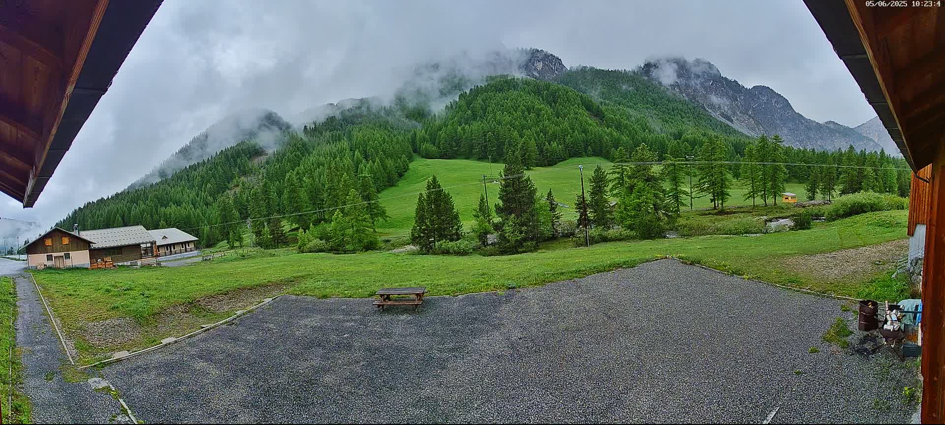A gravel area with a picnic table overlooks a grassy field and a mountainside covered in a dense evergreen forest partially obscured by fog.