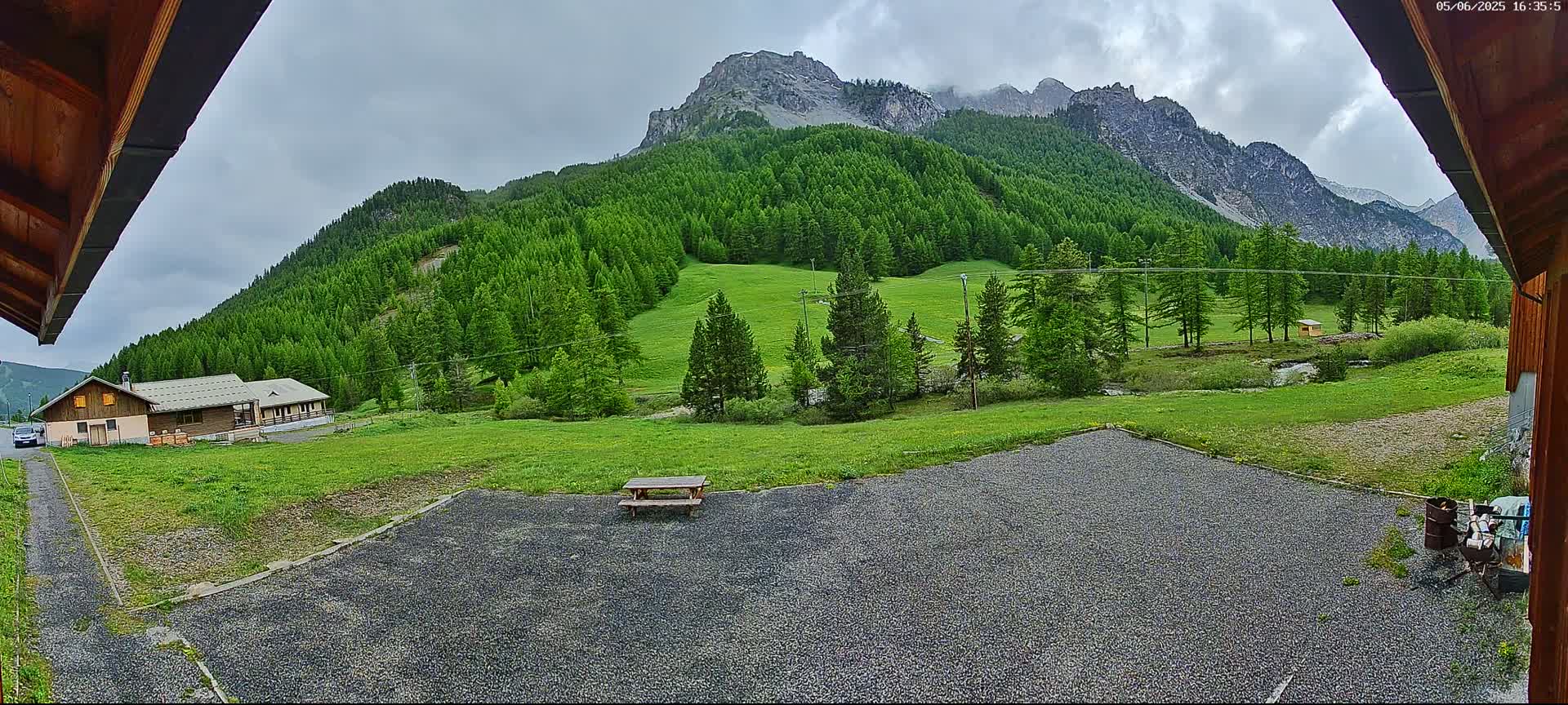 A gravel area with a picnic table sits before a grassy field and a lush green forest covering a mountainside under an overcast sky.