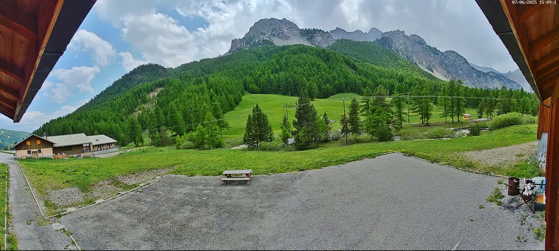 A gravel area with a picnic table sits before a grassy field and a mountain range covered in lush green trees under a partly cloudy sky.