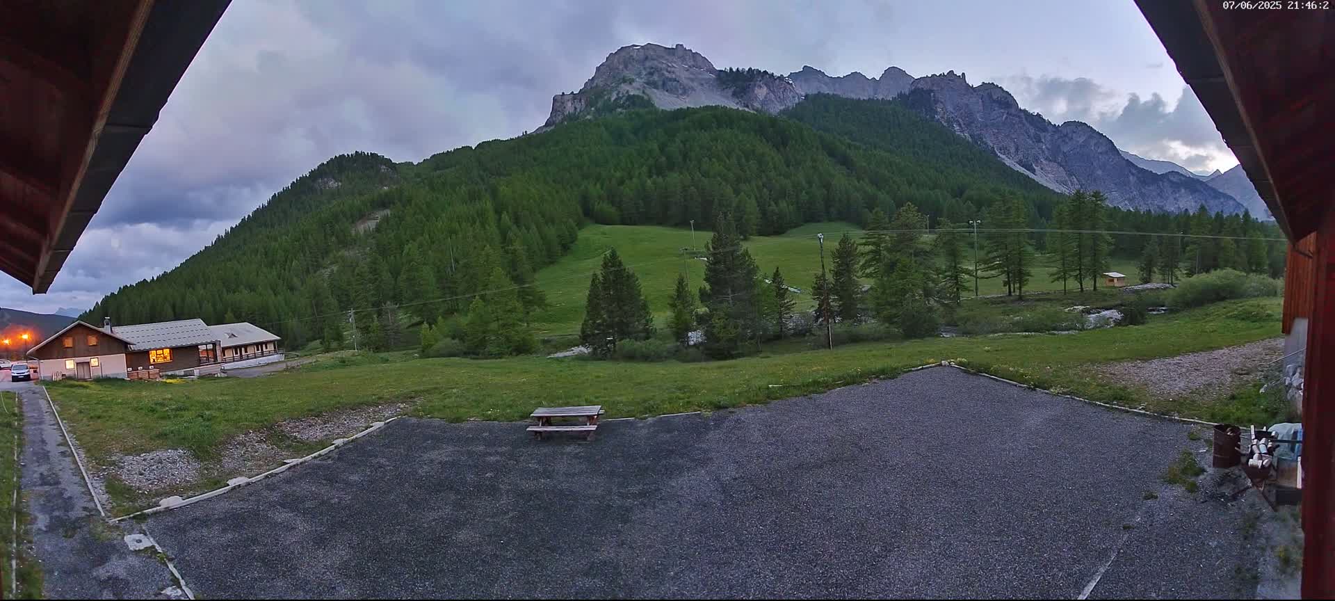 A gravel area with a picnic table sits in front of a building, overlooking a grassy valley and a mountain range under a partly cloudy twilight sky.