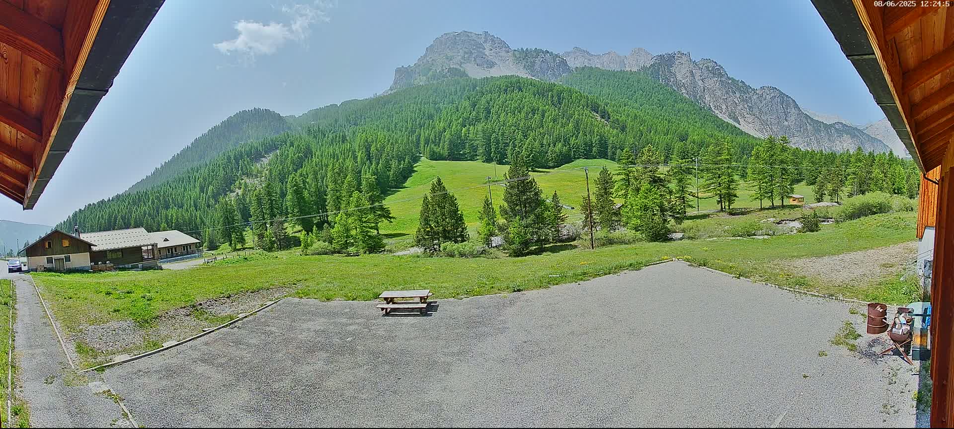 A gravel area with a picnic table overlooks a grassy field and a mountain range covered in a lush green forest under a mostly sunny sky.
