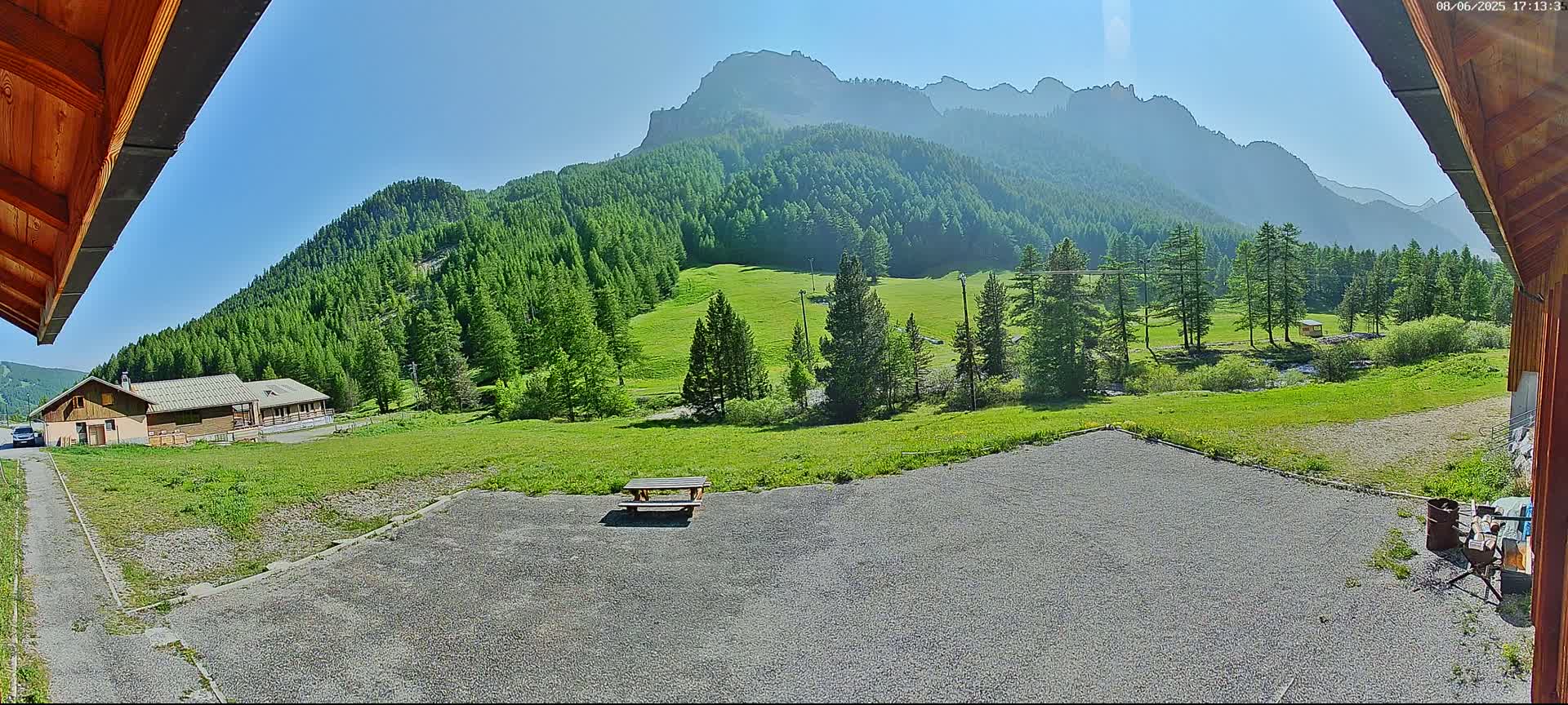 A gravel area with a picnic table sits before a building and expansive green field, all nestled against a backdrop of a mountainous landscape under a clear, sunny sky.