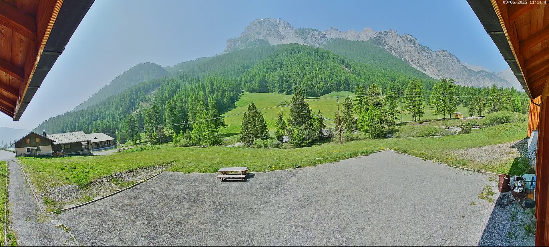 A gravel area with a picnic table sits before a building and a lush green valley, backed by a mountain range under a clear, sunny sky.