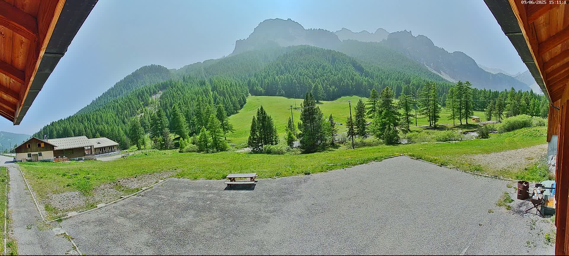 A gravel area with a picnic table overlooks a grassy valley, a cluster of buildings, and a mountain range under a clear, sunny sky.