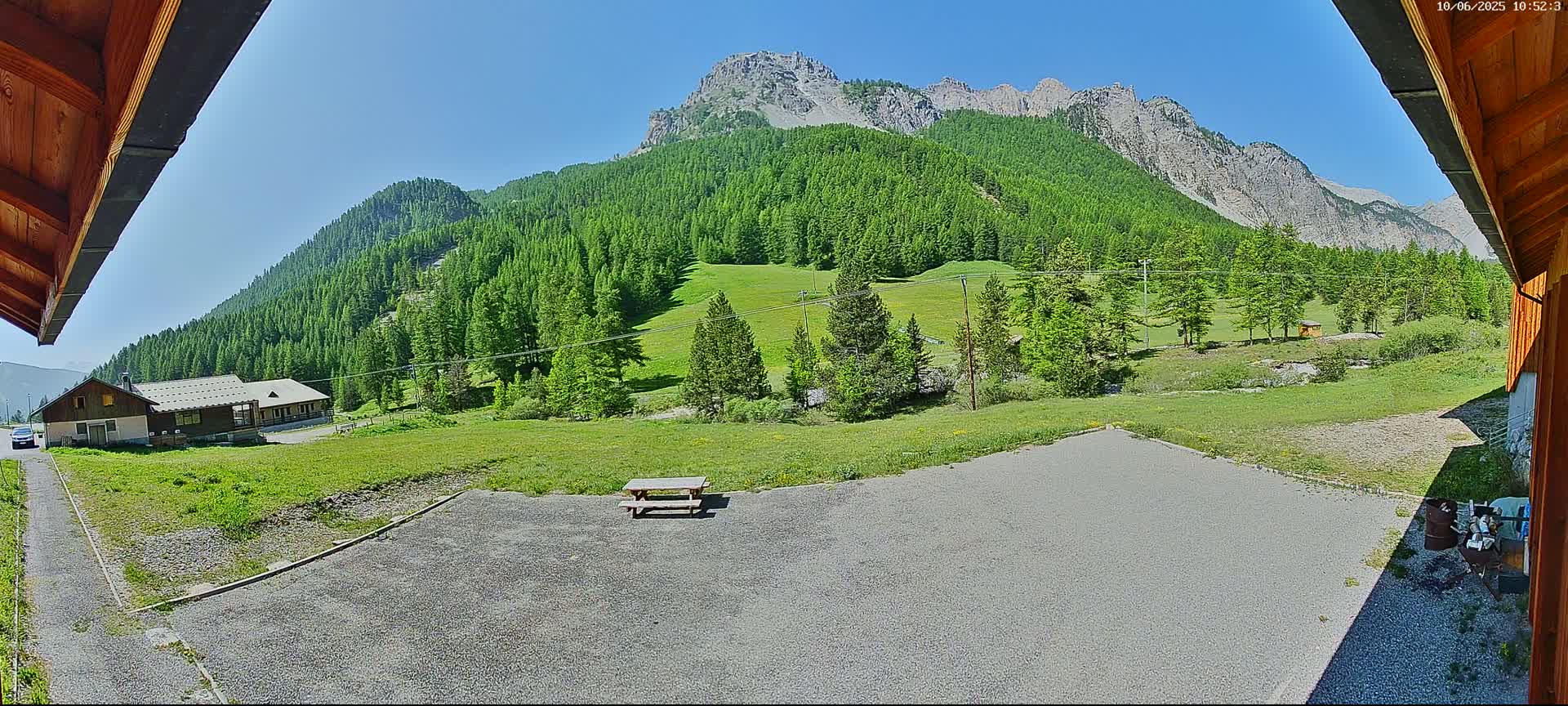 A gravel area with a picnic table sits before a grassy field and a mountain range covered in lush green trees under a sunny sky.