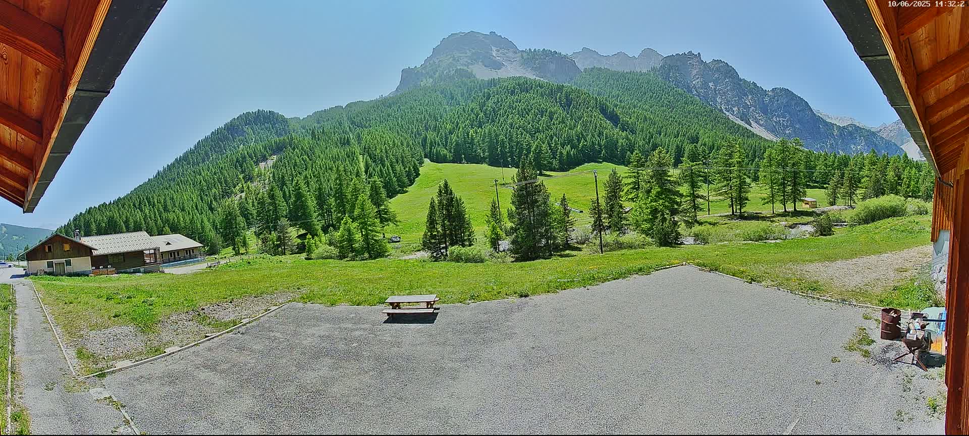 A gravel area with a picnic table sits in front of a building, overlooking a green valley and a mountain range under a clear blue sky.