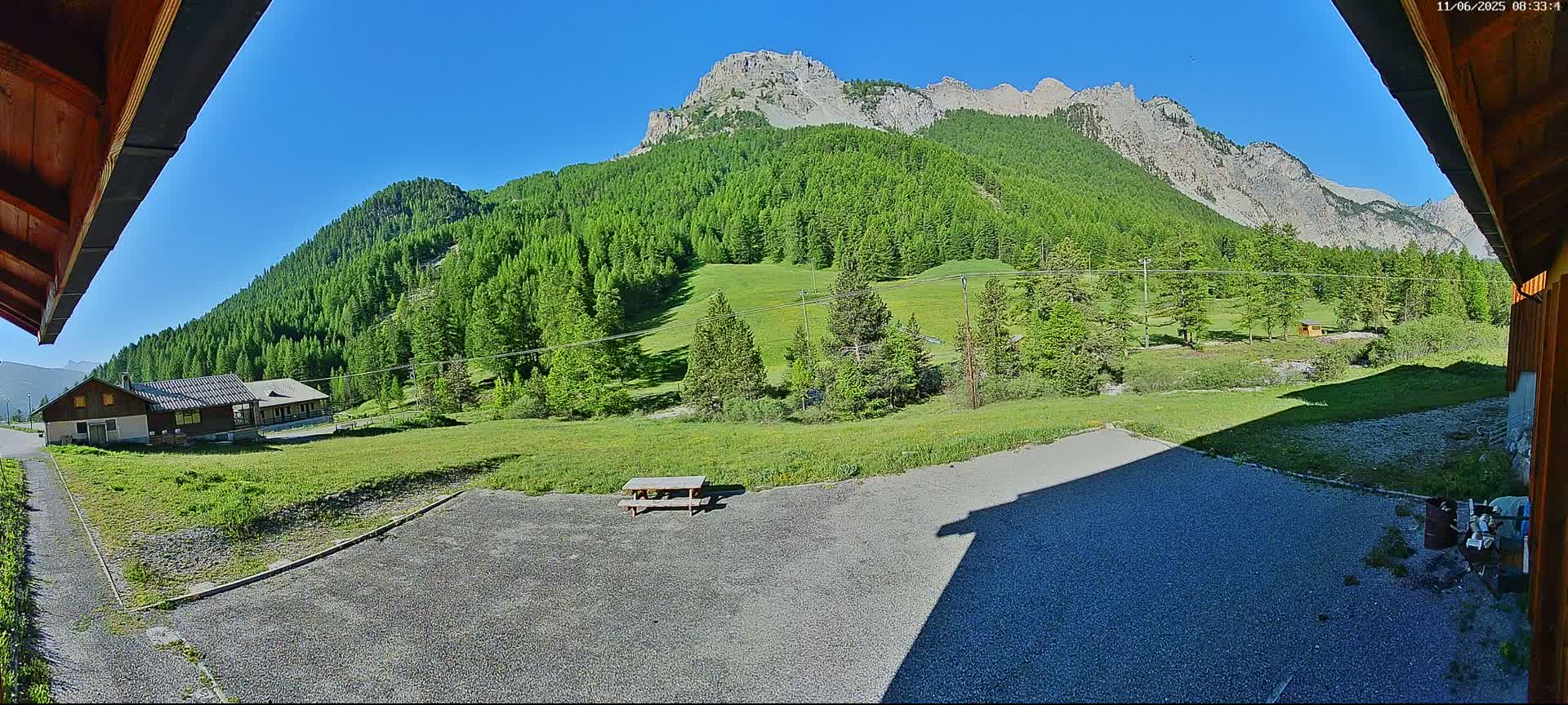 A gravel area with a picnic table sits in front of a small building, overlooking a verdant valley and a mountain range under a clear blue sky.