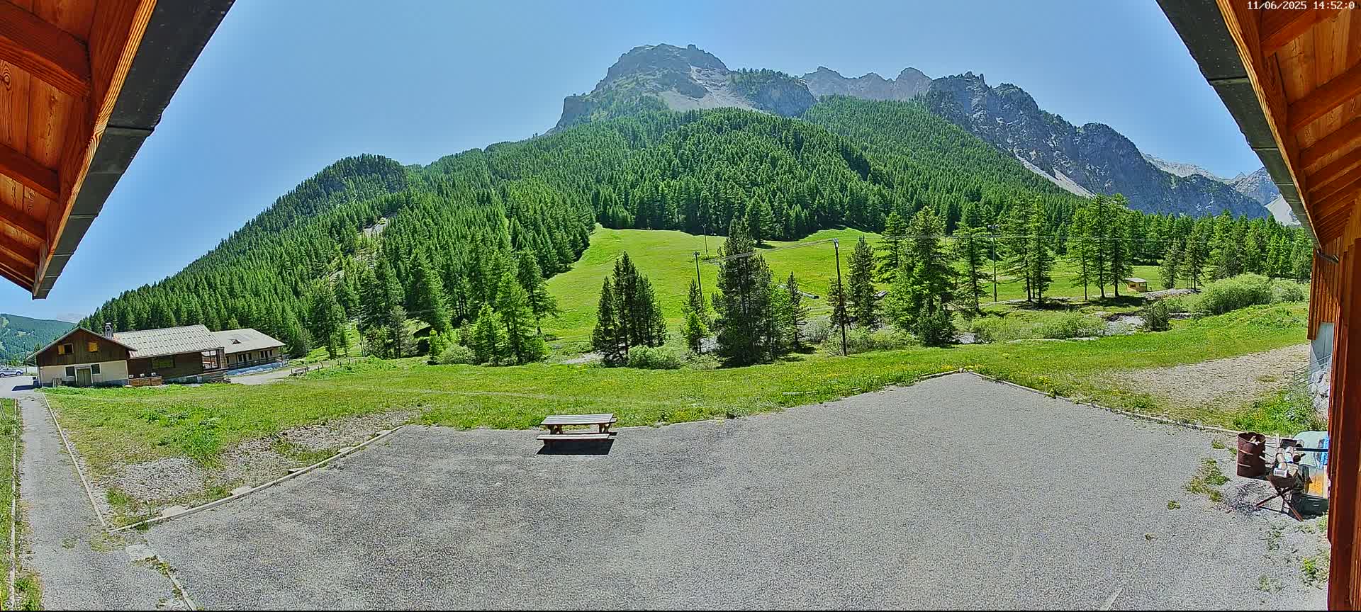 A picnic table sits in a gravel area overlooking a grassy field, a building, and a mountain range under a clear, sunny sky.