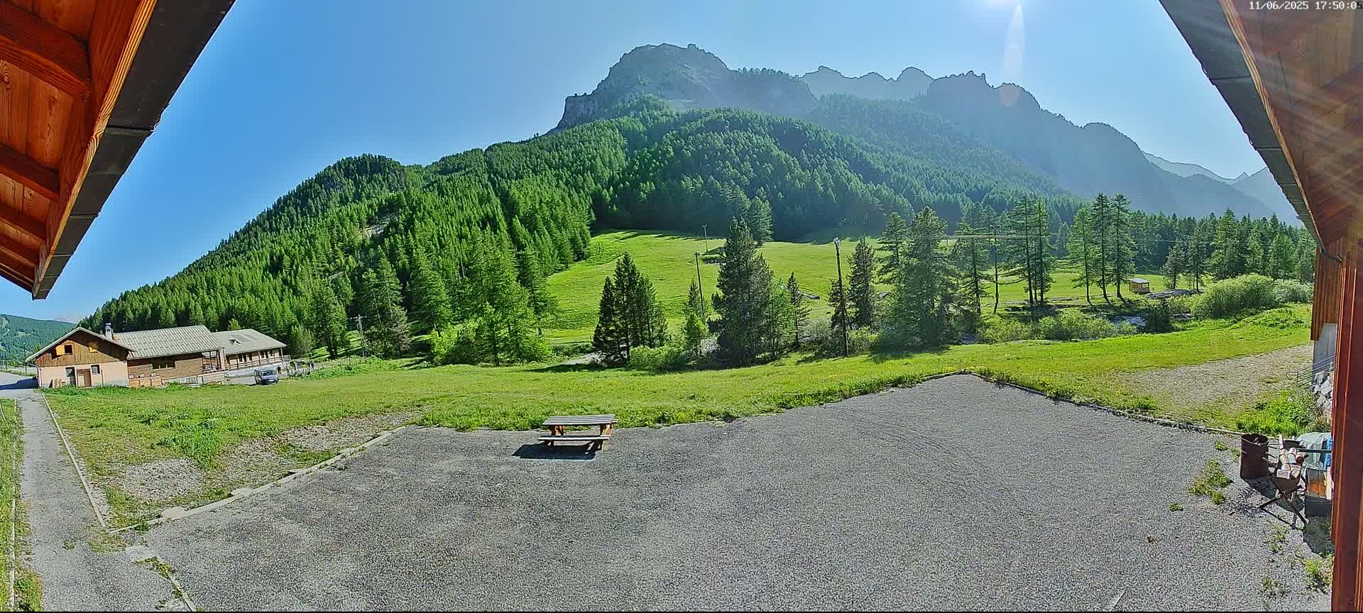 A gravel area with a picnic table sits in front of a building, overlooking a grassy valley and a mountain range under a clear, sunny sky.