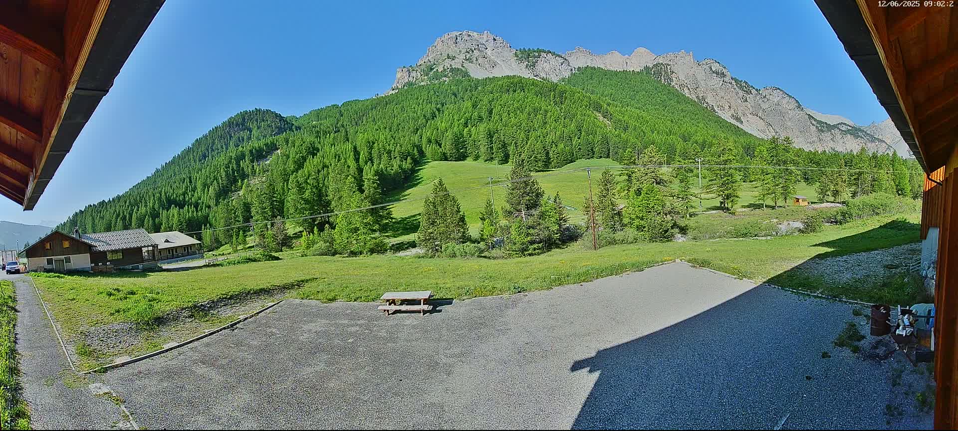 A gravel area with a picnic table sits in front of a building, overlooking a grassy field and a mountain range covered in lush green trees under a clear blue sky.