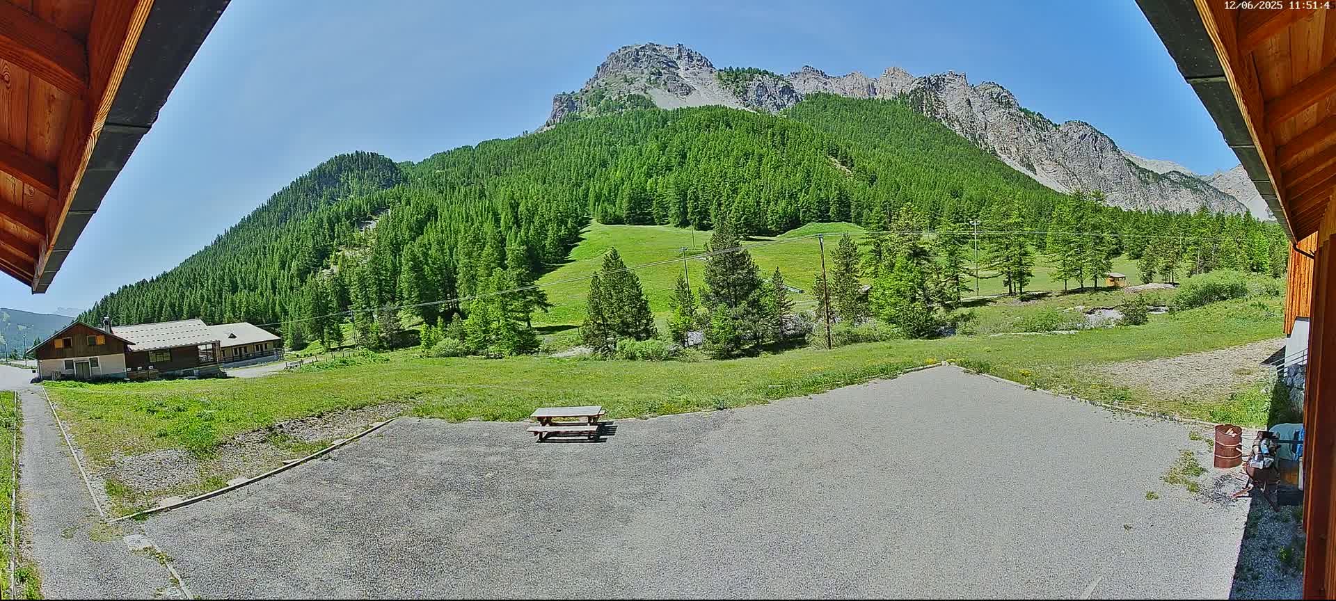 A gravel area with a picnic table overlooks a grassy field and a mountain range covered in lush green pine trees under a clear blue sky.