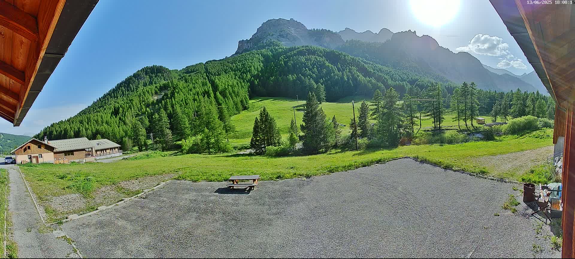 A gravel area with a picnic table sits in front of a building, overlooking a sunny green valley and a mountain range under a mostly clear sky.