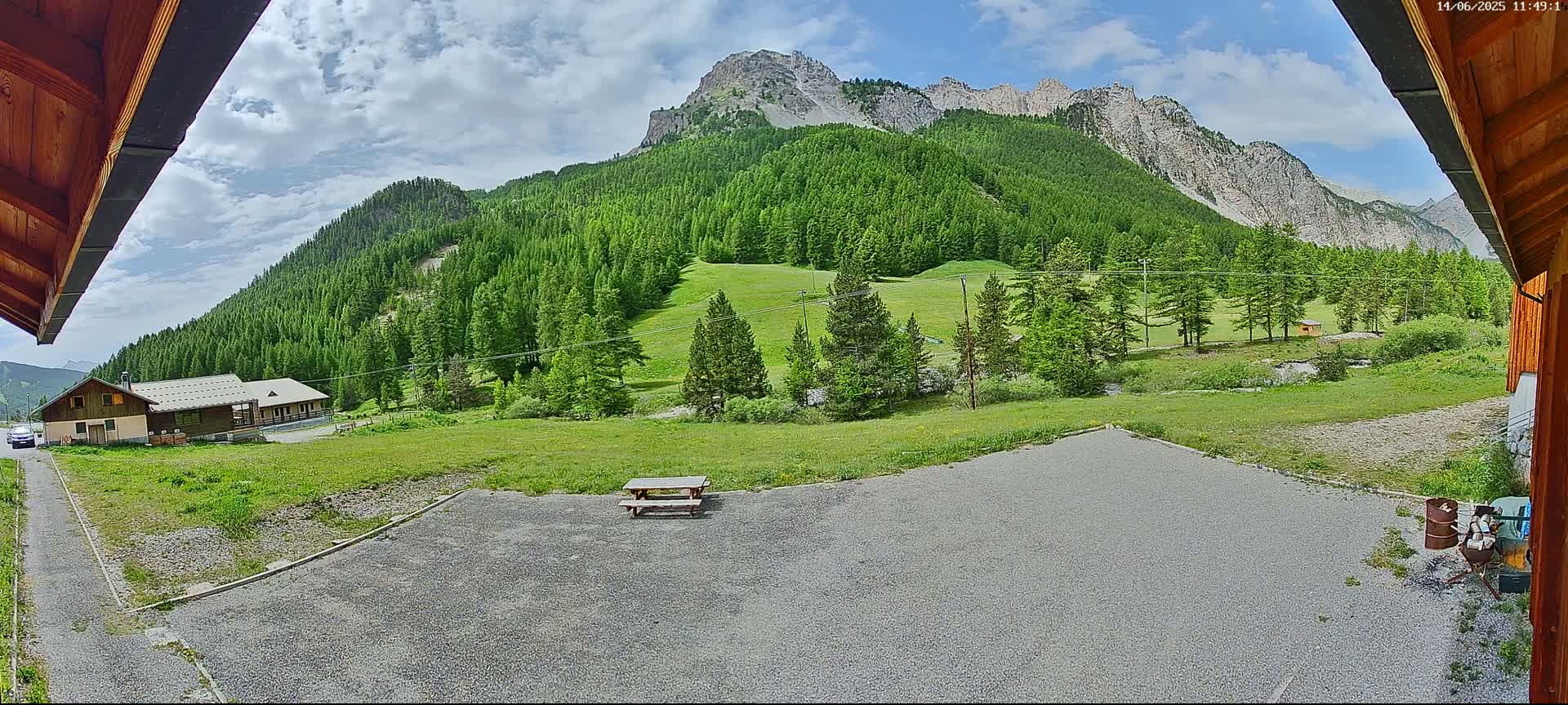 A gravel area with a picnic table sits in front of a building, overlooking a verdant mountainside under a partly cloudy sky.