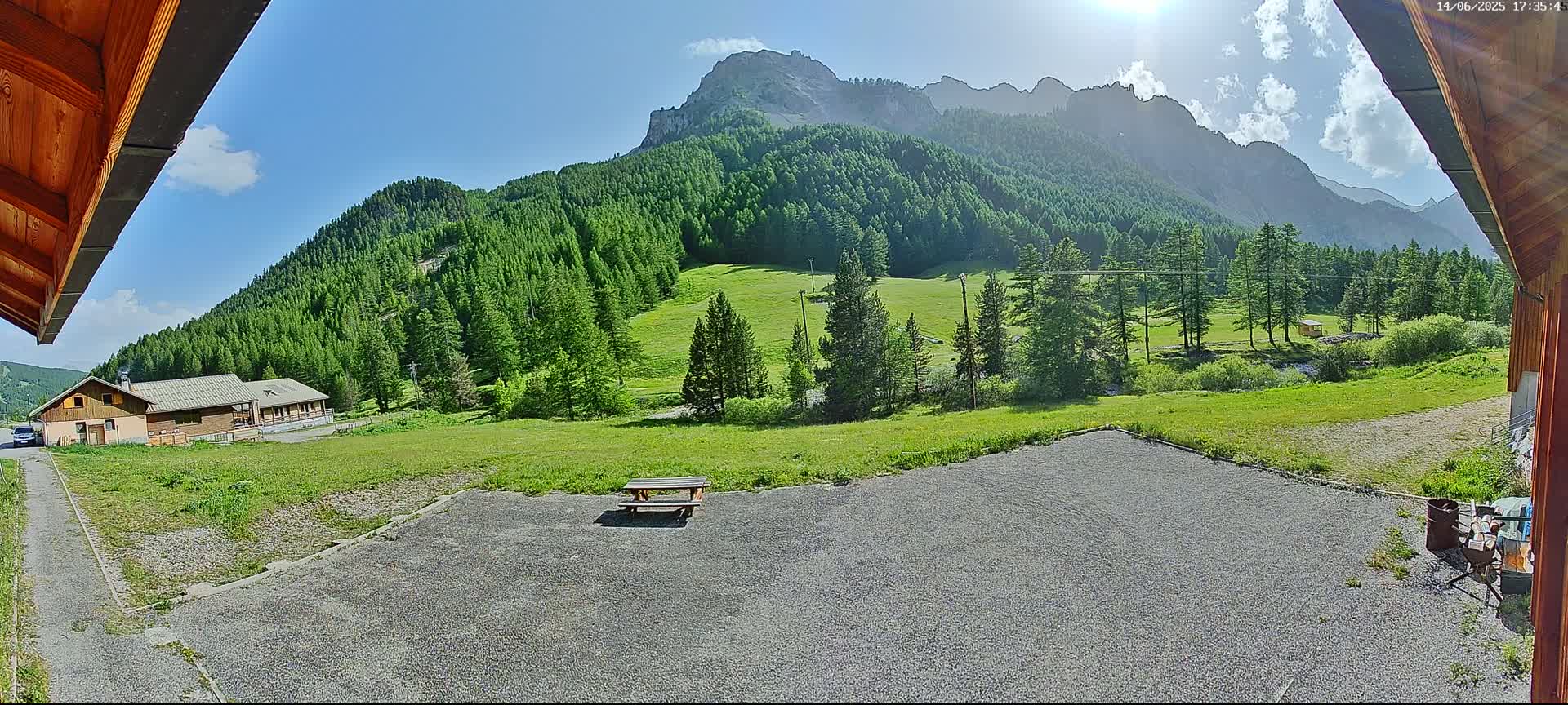 A gravel area with a picnic table sits before a grassy field and a mountainous landscape of lush green forests under a mostly sunny sky.