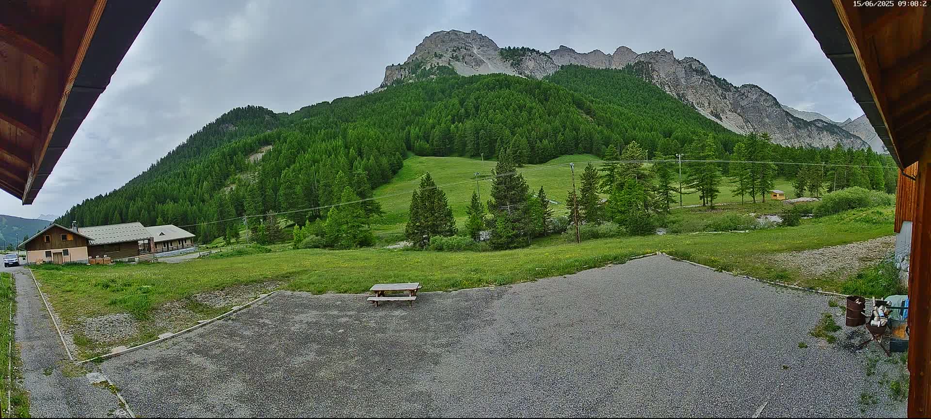 A gravel area with a picnic table sits in front of a building, overlooking a verdant valley and a mountainous backdrop under a cloudy sky.