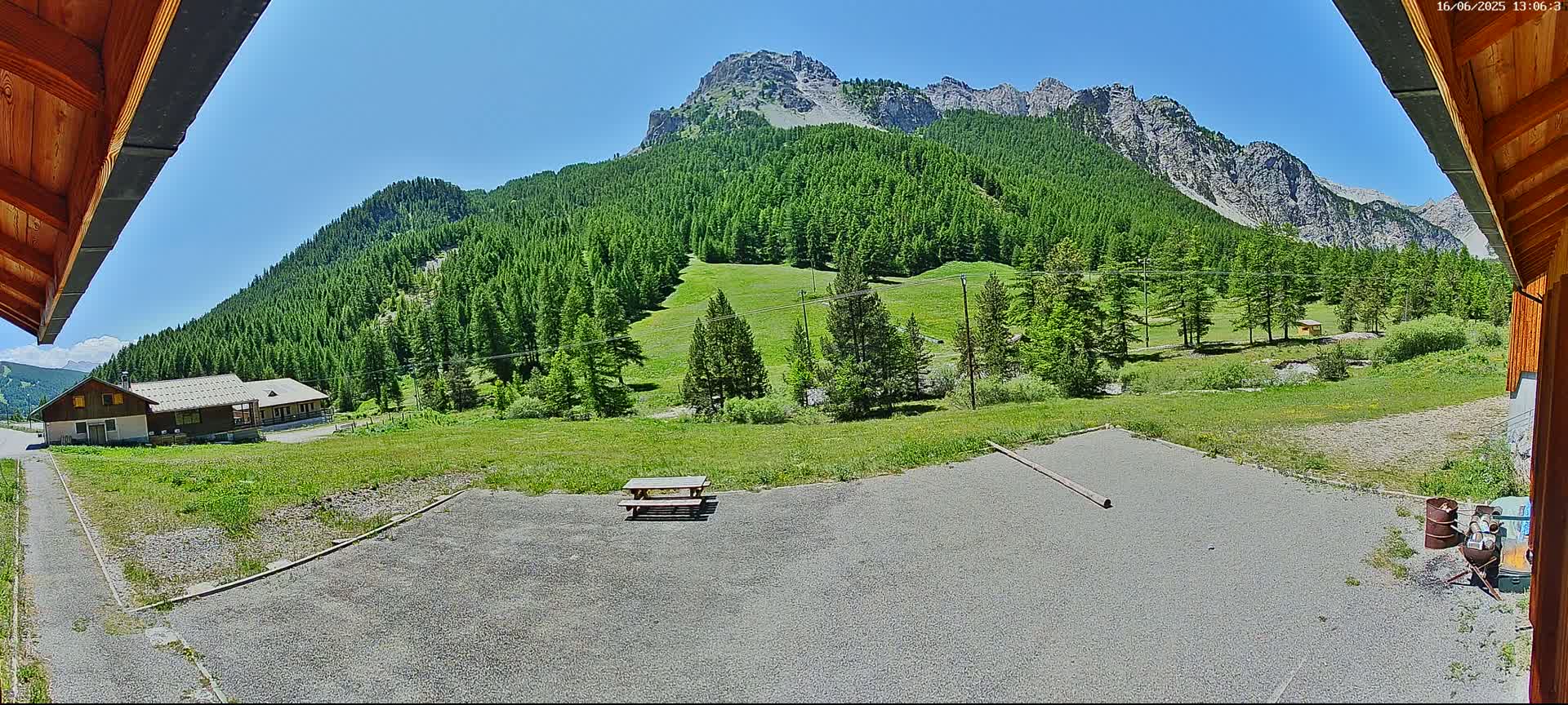 A gravel area with a picnic table sits in front of a building, overlooking a grassy field and a mountain range covered in lush green trees under a clear blue sky.
