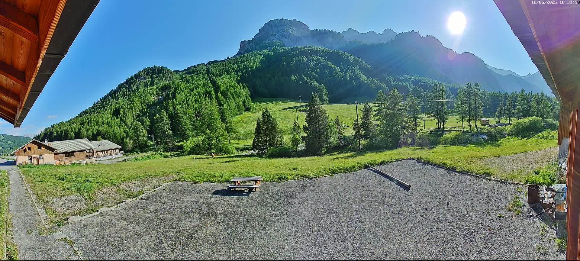 A sunny day reveals a gravel area with a picnic table, a grassy field, a building, and a mountain range covered in lush green trees in the background.