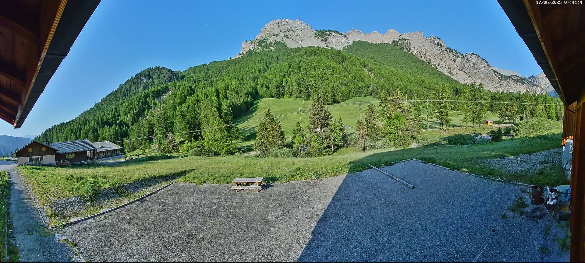 A gravel area with a picnic table sits before a grassy field and a mountainous landscape covered in lush green trees under a clear blue sky.