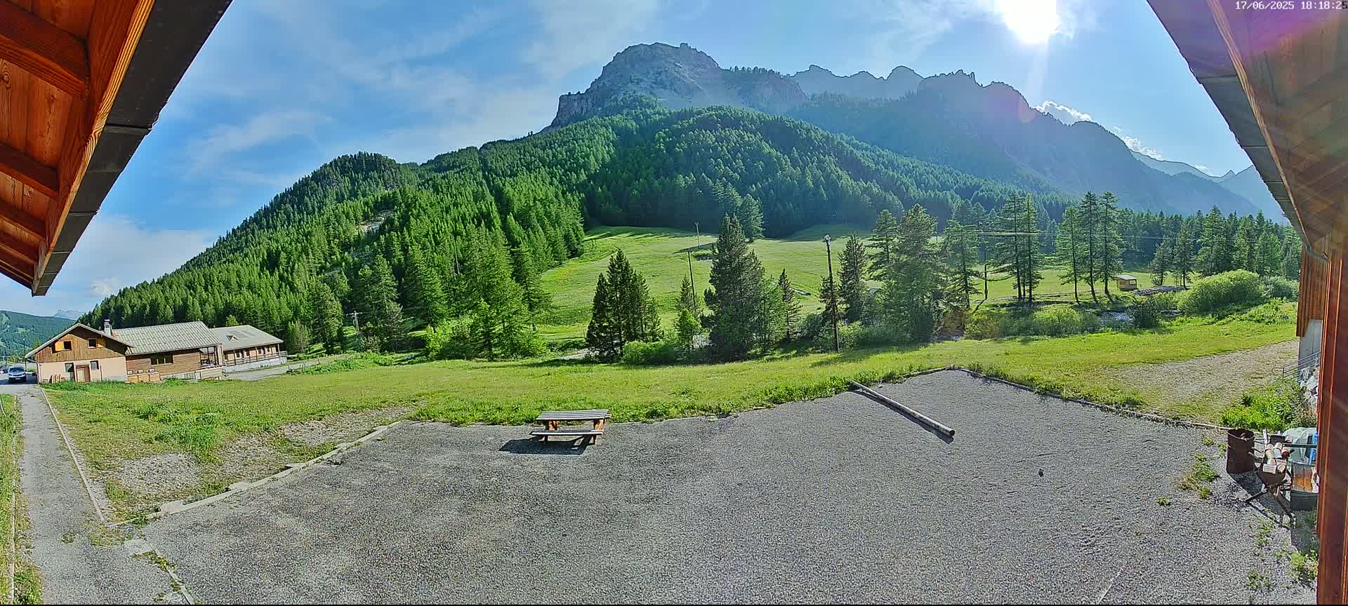A gravel area with a picnic table sits in front of a building, overlooking a sunny valley filled with green meadows and coniferous forests, backed by a mountain range.