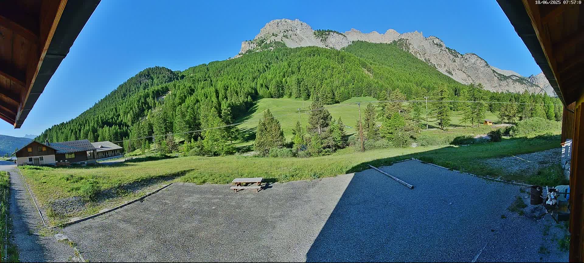 A gravel area with a picnic table sits before a grassy field and a mountain range covered in lush green trees under a clear blue sky.