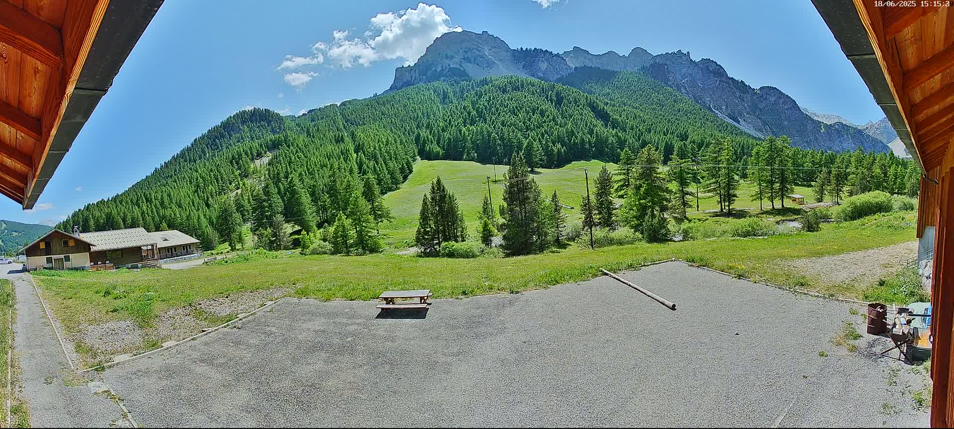 A gravel area with a picnic table is in the foreground, overlooking a grassy field, a building, and a mountainous landscape under a mostly sunny sky.