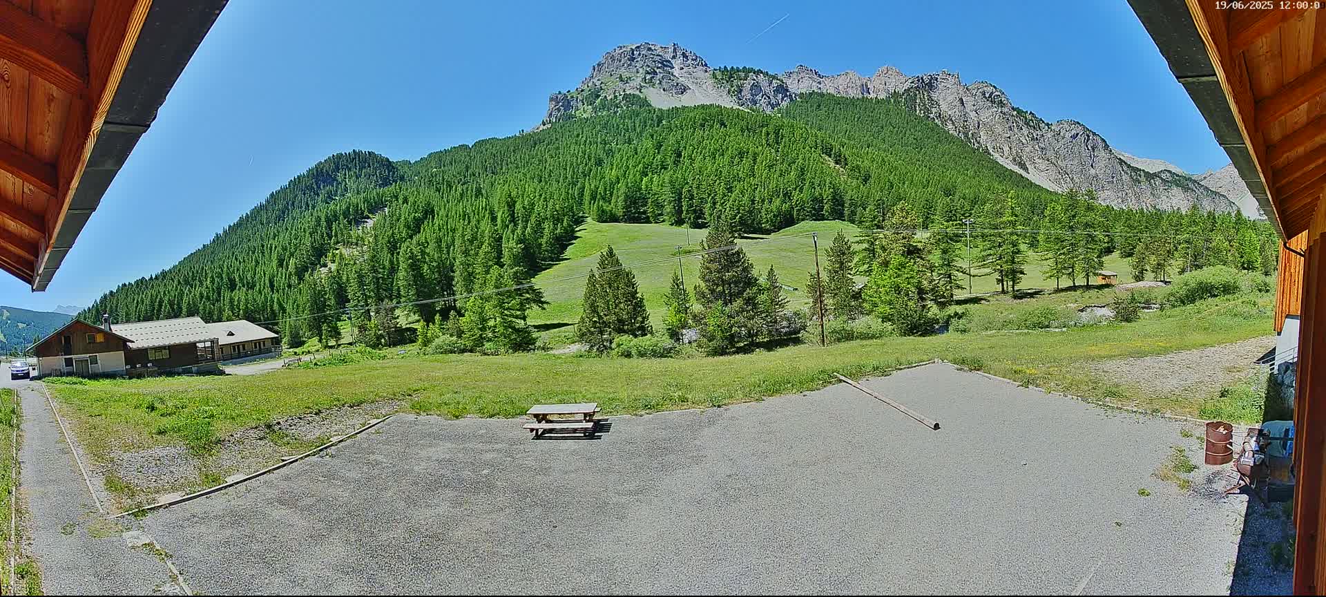 A gravel area with a picnic table sits in front of a building, overlooking a lush green valley and mountains under a clear blue sky.
