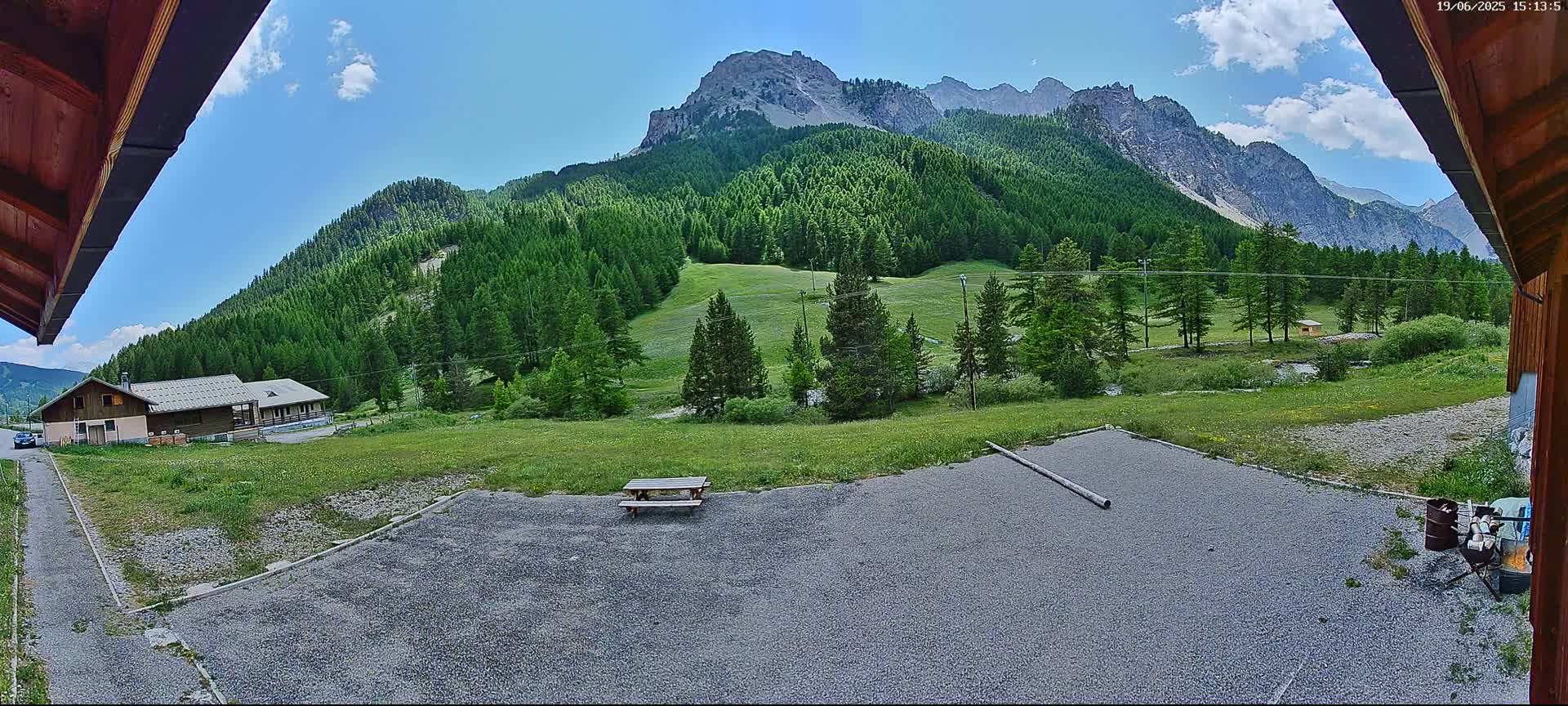 A gravel area with a picnic table is in the foreground, overlooking a valley of green trees and mountains under a mostly sunny sky.