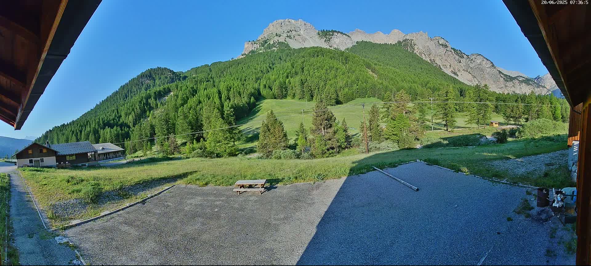 A gravel area with a picnic table sits in front of a building, overlooking a grassy field, a forest of evergreen trees, and a mountain range under a clear blue sky.