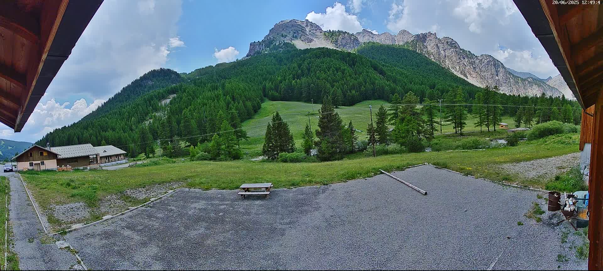 A gravel area with a picnic table sits before a grassy field and a mountain range covered in lush green trees under a partly cloudy sky.