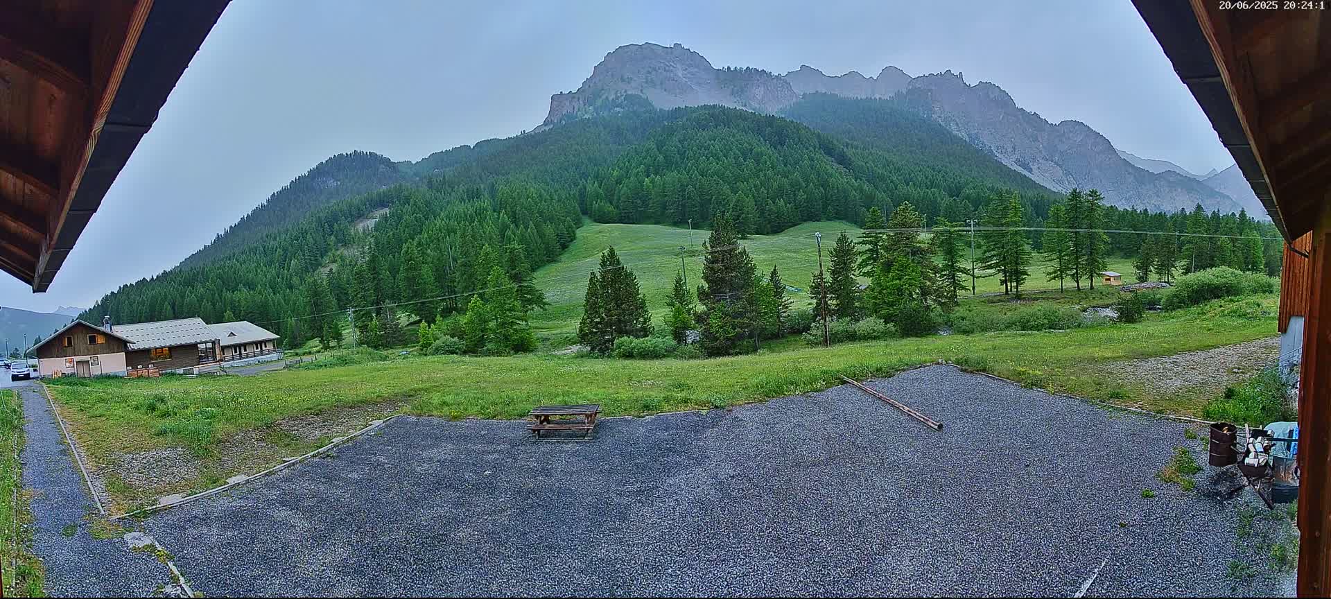 A gravel area with a picnic table is situated in front of a building, overlooking a verdant valley and mountains under an overcast sky.
