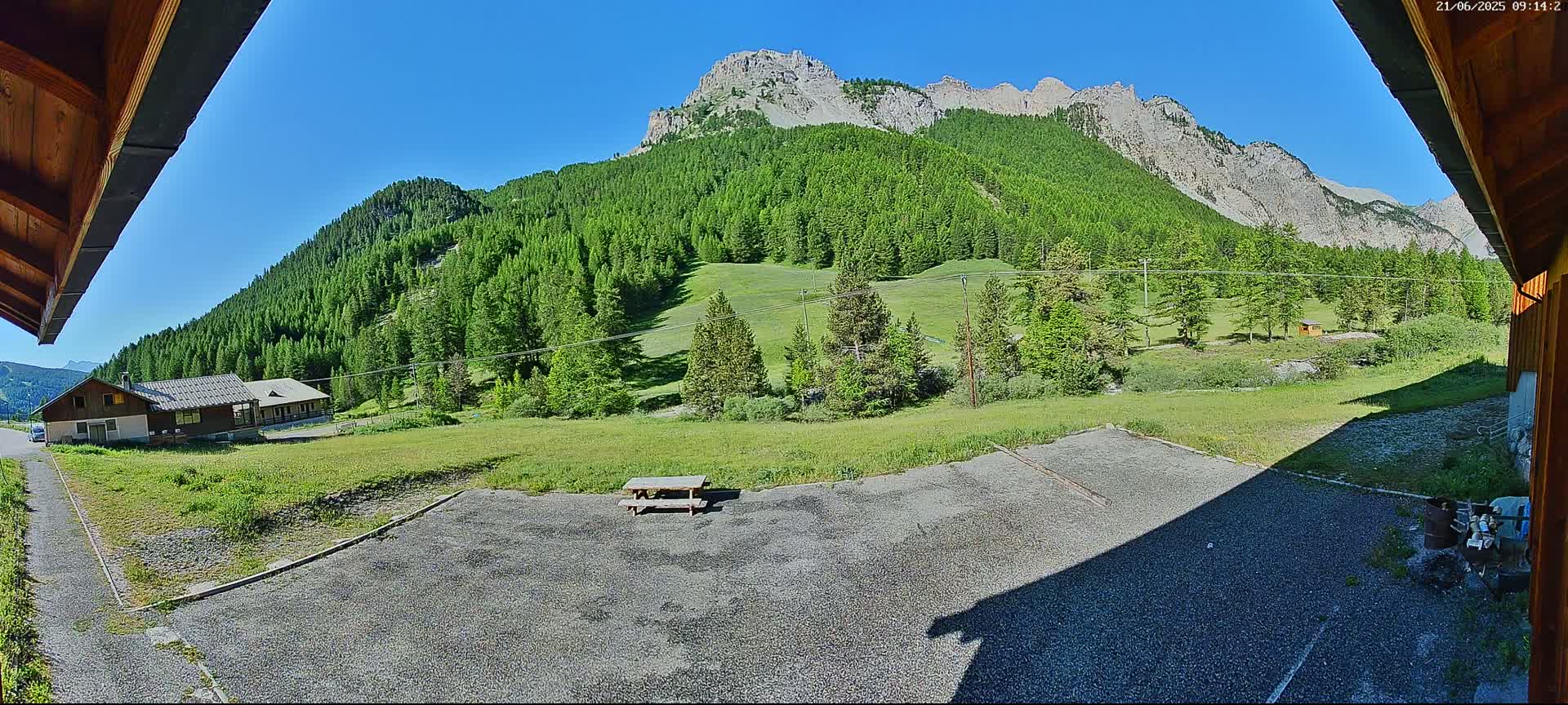 A gravel area with a picnic table sits in front of a building, overlooking a verdant valley and a mountain range under a clear blue sky.