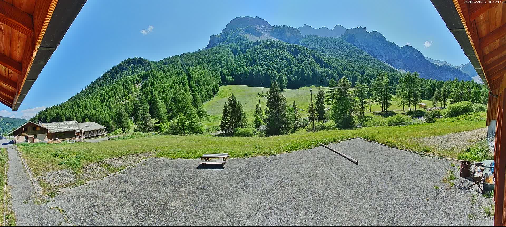 A picnic table sits on a gravel area overlooking a grassy field, a building, and a mountainous landscape under a clear, sunny sky.