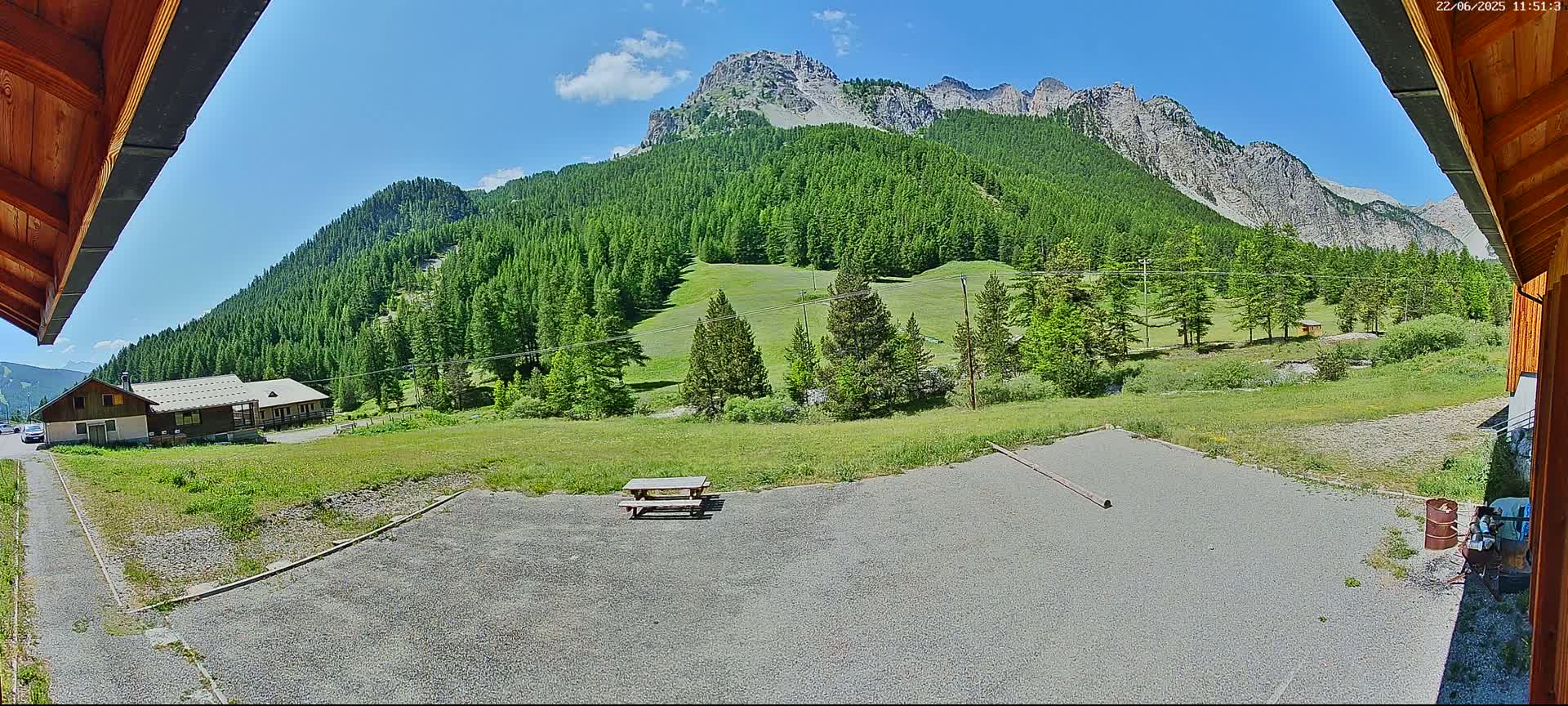 A gravel area with a picnic table sits before a grassy field and a large mountain covered in lush green trees under a mostly sunny sky.
