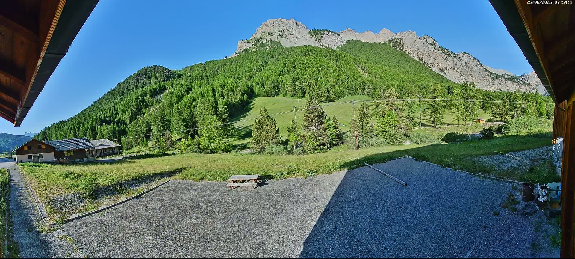 A gravel area with a picnic table sits in front of a building, overlooking a grassy field and a mountain range covered in lush green trees under a clear blue sky.