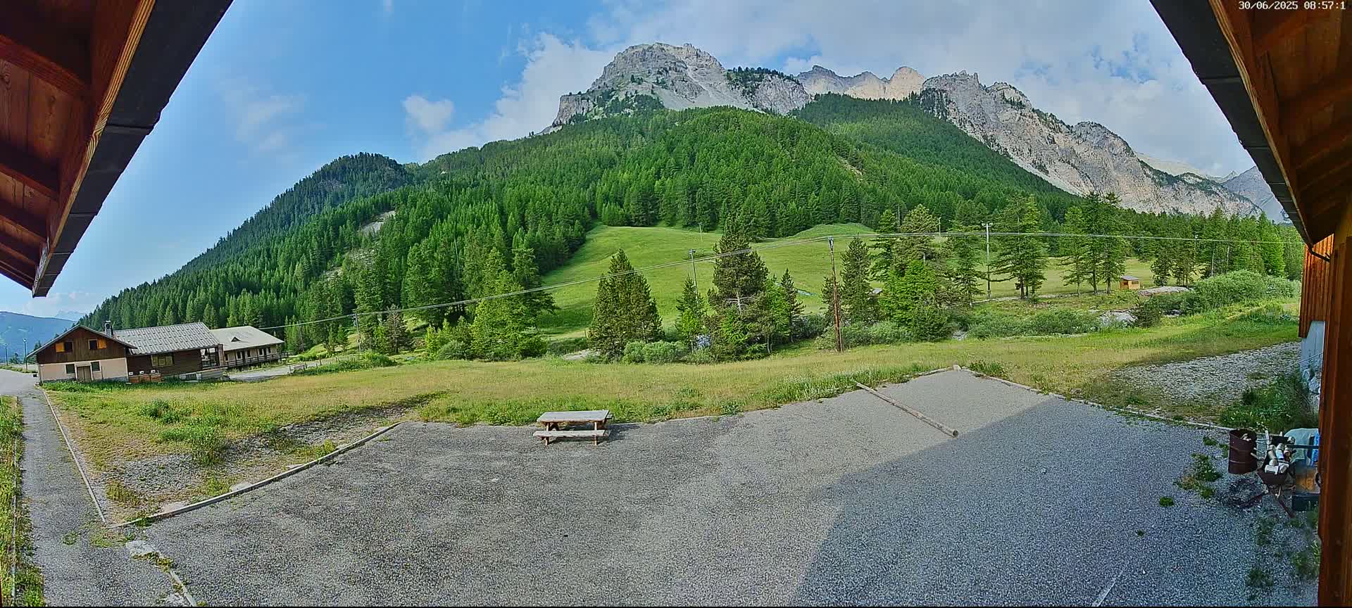 A gravel area with a picnic table sits in front of a building, overlooking a verdant valley and a towering mountain range under a partly cloudy sky.