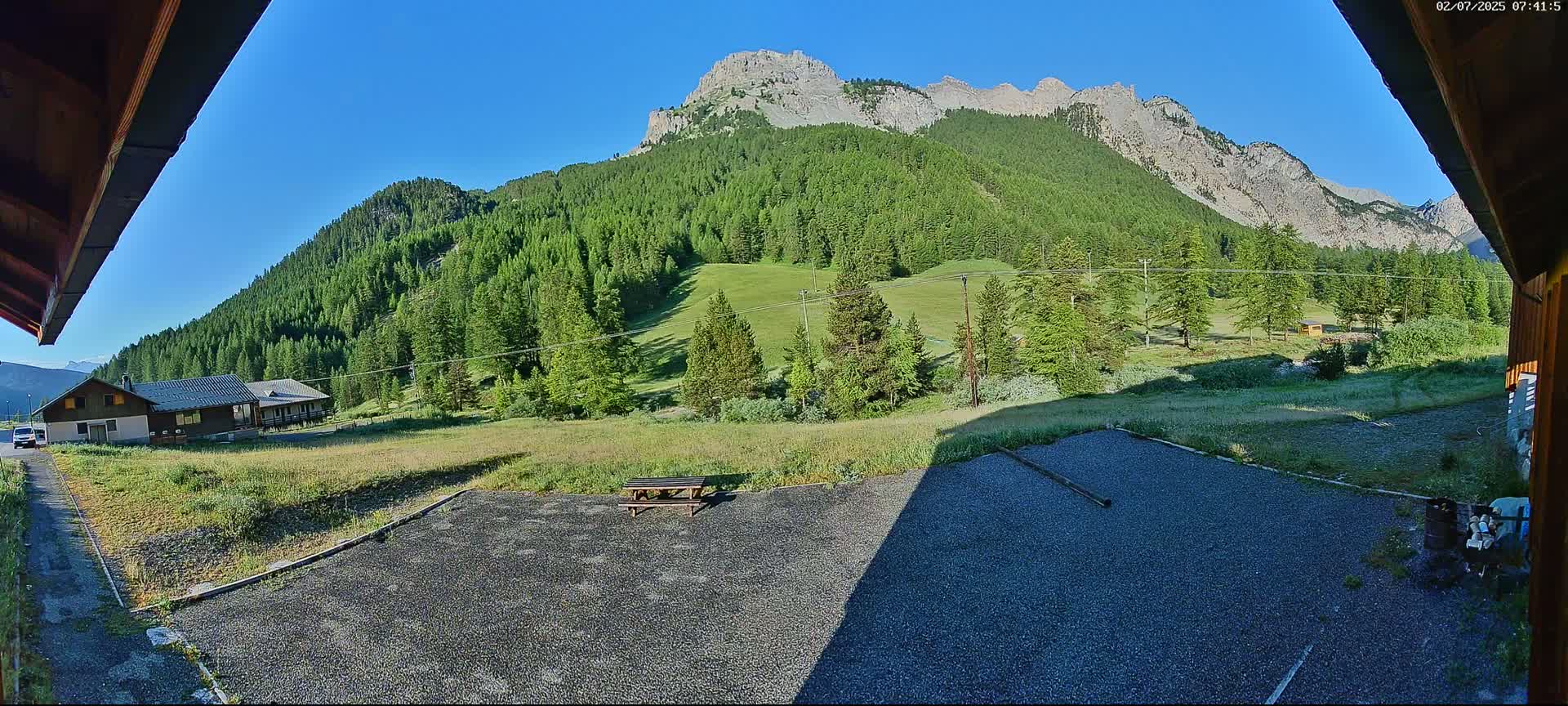 A gravel area with a picnic table sits in front of a building, overlooking a valley of green trees and mountains under a clear blue sky.