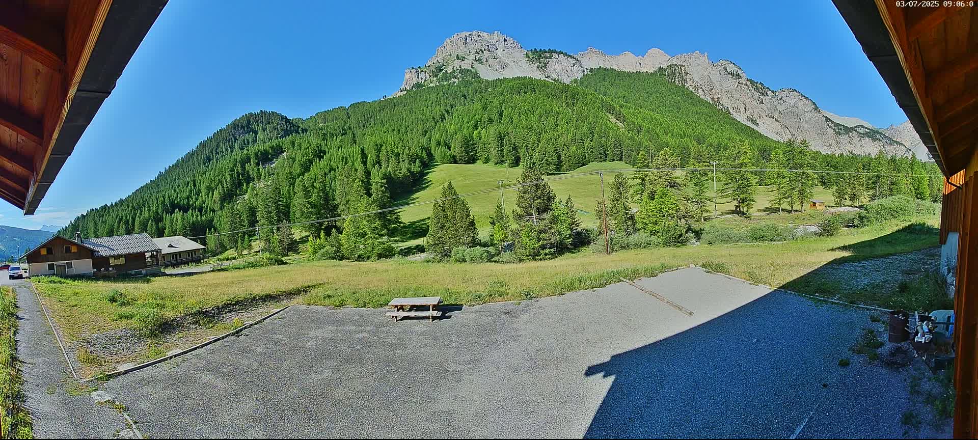 A gravel area with a picnic table is situated in front of a building, overlooking a green valley and a large mountain range under a clear blue sky.